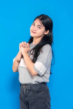 Umut dolu bir ifadeyle genç Asyalı kadın, elleri birbirine kenetlenmiş, nazikçe gülümsüyor. Minnettar ve heyecanlı görünüyor, canlı mavi bir arka plana karşı iyimserliğini ifade ediyor..