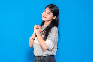 Umut dolu bir ifadeyle genç Asyalı kadın, elleri birbirine kenetlenmiş, nazikçe gülümsüyor. Minnettar ve heyecanlı görünüyor, canlı mavi bir arka plana karşı iyimserliğini ifade ediyor..