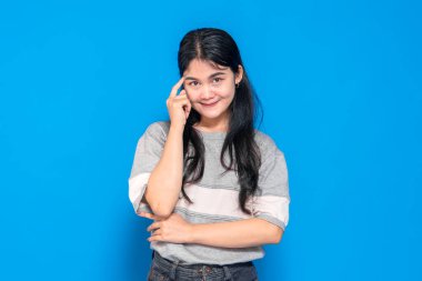 Kendine güvenen ve düşünceli bir ifadeyle genç Asyalı kadın şakağına hafifçe gülümsüyor. Zeki ve yaratıcı görünüyor, parlak mavi bir arka plana karşı duruyor..