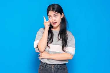 Meraklı ve şaşırmış bir ifadeyle genç bir Asyalı kadın işaret parmağını bir fikri varmış gibi kaldırıyor. O düşünceli ve ilham verici görünüyor, mavi bir arka planda duruyor..