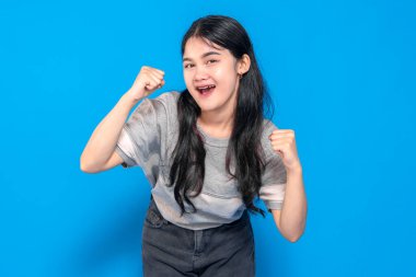 Uzun siyah saçlı, gri tişörtlü, neşeli bir gülümsemeyle heyecanını ifade eden ve sağlam mavi bir arka plan karşısında yumruklarını sıkan Asyalı genç bir kadın..