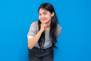 Uzun siyah saçlı Asyalı genç bir kadın, gri bir gömlek giyiyor, parmaklarıyla dudaklarında şşşş hareketi yapıyor, gizemli bir şekilde mavi bir arka plana gülümsüyor..