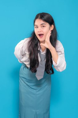 Üniformalı Endonezyalı bir lise öğrencisi eli ağzının yanındayken şakacı bir şekilde bağırıyor. Heyecan ve neşe ifade ediyor, canlı mavi bir arka plana karşı neşeli görünüyor..