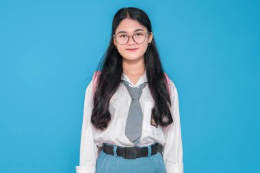 Endonezyalı bir lise öğrencisi gözlüklü, beyaz üniformalı ve gri kravatlı, hafif bir gülümseme ve mavi bir arka planda duran bir sırt çantasıyla..