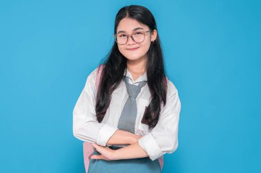 Beyaz üniformalı, mavi etekli, gözlüklü, pembe sırt çantalı, kolları çapraz ve mavi arka planda hafifçe gülümseyen Endonezyalı bir liseli kız..
