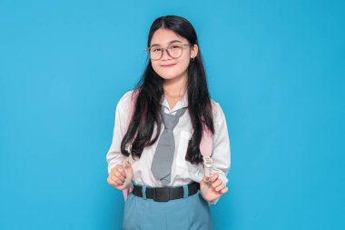 Endonezyalı liseli bir kız, gözlüklü, beyaz üniformalı ve mavi etekli, pembe sırt çantasıyla mavi arka planda duran, güvenle gülümseyen..