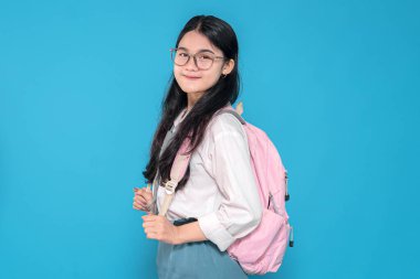 Endonezyalı liseli bir kız, gözlüklü, beyaz üniformalı ve mavi etekli, pembe sırt çantasıyla mavi arka planda duran, güvenle gülümseyen..
