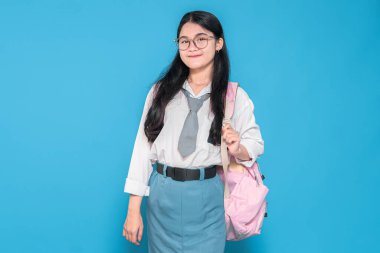 Beyaz üniformalı, mavi etekli, gözlüklü ve kravatlı, pembe sırt çantasıyla mavi arka planda duran Endonezyalı bir liseli kız..