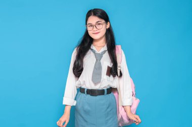 Beyaz üniformalı, mavi etekli, gözlüklü ve kravatlı, pembe sırt çantasıyla mavi arka planda duran Endonezyalı bir liseli kız..