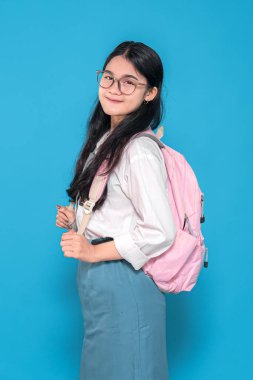 Endonezyalı liseli bir kız, gözlüklü, beyaz üniformalı ve mavi etekli, pembe sırt çantasıyla mavi arka planda duran, güvenle gülümseyen..