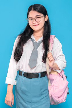 Beyaz üniformalı, mavi etekli, gözlüklü ve kravatlı, pembe sırt çantasıyla mavi arka planda duran Endonezyalı bir liseli kız..