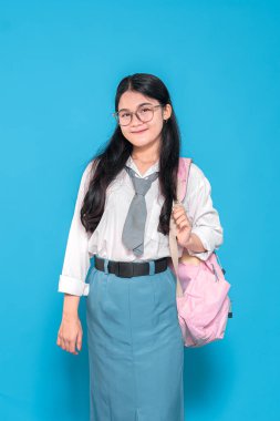 Beyaz üniformalı, mavi etekli, gözlüklü ve kravatlı, pembe sırt çantasıyla mavi arka planda duran Endonezyalı bir liseli kız..