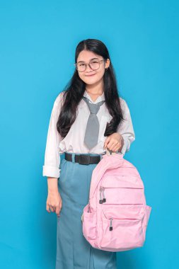 Beyaz üniformalı ve mavi etekli Endonezyalı bir liseli kız, gözlük takıyor, pembe bir sırt çantası taşıyor, mavi arka planda düşünceli bir gülümsemeyle yan tarafa bakıyor..