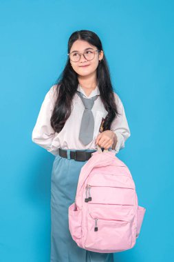 Beyaz üniformalı ve mavi etekli Endonezyalı bir liseli kız, gözlük takıyor, pembe bir sırt çantası taşıyor, mavi arka planda düşünceli bir gülümsemeyle yan tarafa bakıyor..