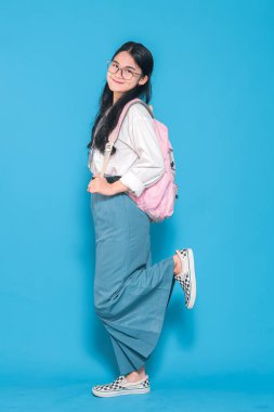 Beyaz üniformalı, mavi etekli, gözlüklü, pembe çantalı bir Endonezyalı liseli kız. Bir bacağı havada, mavi arka planda neşeli bir şekilde poz veriyor..