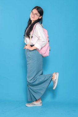 Beyaz üniformalı, mavi etekli, gözlüklü, pembe çantalı bir Endonezyalı liseli kız. Bir bacağı havada, mavi arka planda neşeli bir şekilde poz veriyor..