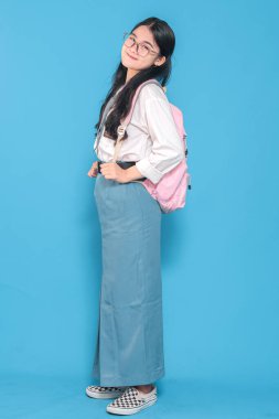 Beyaz üniformalı, mavi etekli, kravat ve gözlük takan, pembe sırt çantasıyla güvenle gülümseyen Endonezyalı bir liseli kız..