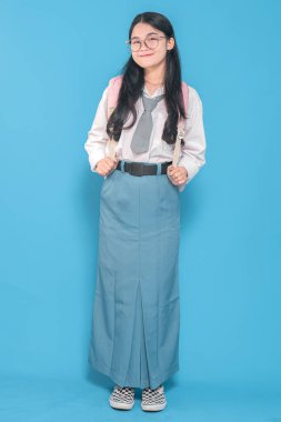 Beyaz üniformalı, mavi etekli, kravat ve gözlük takan, pembe sırt çantasıyla güvenle gülümseyen Endonezyalı bir liseli kız..