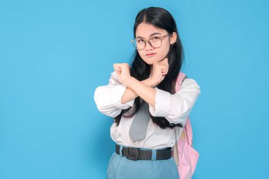 Beyaz üniformalı, mavi etekli, gözlüklü ve kravatlı, pembe bir sırt çantası taşıyan, mavi arka planda kararlı bir ifadeyle X işaretinde kollarını birleştiren Endonezyalı bir liseli kız..