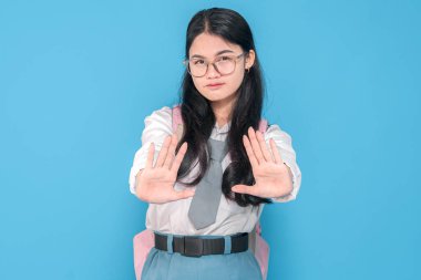 Beyaz üniformalı, mavi etekli, gözlüklü ve kravatlı, pembe bir sırt çantası olan Endonezyalı bir liseli kız iki eliyle bir dur hareketi yapıyor ve mavi arka planda ciddi bir ifade takınıyor..