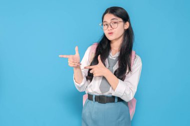 Beyaz üniformalı, mavi etekli, gözlüklü ve pembe sırt çantalı Endonezyalı bir liseli kız, mavi arka planda somurtkan bir ifadeyle şakayla parmak tabancası hareketi yapıyor..