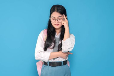 Beyaz üniformalı, mavi etekli, gözlüklü ve pembe sırt çantalı Endonezyalı bir liseli kız mavi arka planda elini şakağına koyup düşünceli bir şekilde poz veriyor..