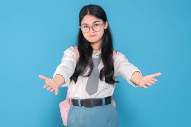 Beyaz üniformalı, mavi etekli, gözlüklü ve pembe sırt çantalı Endonezyalı bir liseli kız mavi bir arka plana karşı kollarını öne uzatarak yalvarıyor..