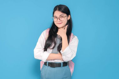 Beyaz üniformalı, mavi etekli, gözlüklü ve pembe sırt çantalı Endonezyalı bir liseli kız mavi arka plana karşı güvenle gülümsüyor..