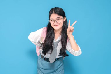 Beyaz üniformalı ve mavi etekli Endonezyalı bir liseli kız barış işareti yaparken neşeyle gülümsüyor. Gözlük takıyor ve mavi arka planda pembe bir sırt çantası taşıyor..