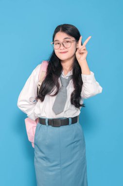 Endonezyalı liseli bir kız, gözlük takıyor, beyaz üniformalı ve mavi etekli. Mavi arka planda parmaklarıyla barış işareti yaparken kendine güvenen bir şekilde gülümsüyor..