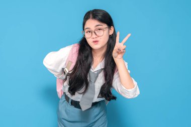 Beyaz üniformalı ve mavi etekli Endonezyalı bir liseli kız barış işareti yaparken neşeyle gülümsüyor. Gözlük takıyor ve mavi arka planda pembe bir sırt çantası taşıyor..