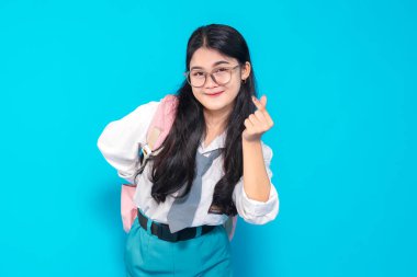 Endonezyalı bir lise öğrencisi, uzun siyah saçlı, gözlüklü ve okul üniformalı. Parlak mavi bir arkaplan karşısında neşeli bir ifadeyle parmak kalp hareketi yapıyor..