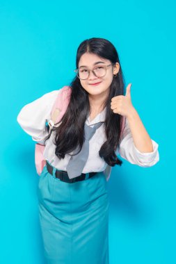 Uzun siyah saçlı, gözlüklü, beyaz bluzlu ve mavi etekli Endonezyalı bir lise öğrencisi mavi arka planda güvenle gülümsüyor..