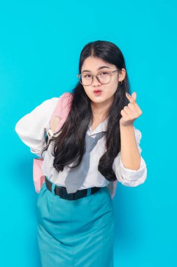 Endonezyalı bir lise öğrencisi, uzun siyah saçlı, gözlüklü ve okul üniformalı. Parlak mavi bir arkaplan karşısında neşeli bir ifadeyle parmak kalp hareketi yapıyor..