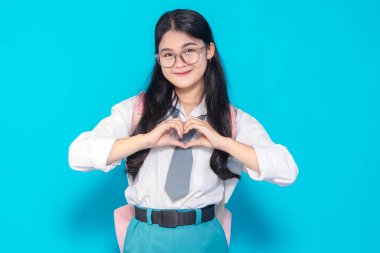 Endonezyalı bir lise öğrencisi, okul üniforması içinde, elleriyle kalp şeklini oluştururken sıcak bir şekilde gülümsüyor. Parlak mavi bir arka planda duran bir sırt çantası ve gözlük takıyor..
