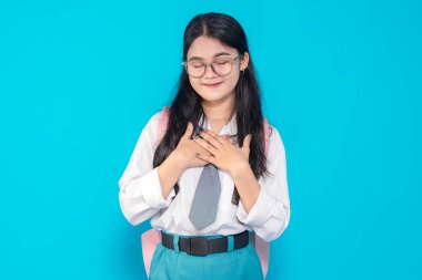 Endonezyalı bir lise öğrencisi, gözlüklü ve sırt çantalı, gözleri kapalı, elleri göğsünde gülümseyen parlak mavi bir geçmişe karşı şükranlarını sunuyor..