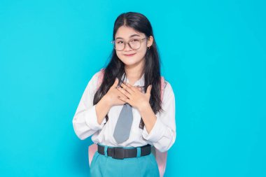 Endonezyalı bir lise öğrencisi, gözlüklü, okul üniformalı ve sırt çantalı ellerini göğsüne koyup gülümseyen parlak mavi bir geçmişe karşı şükranlarını sunuyor..