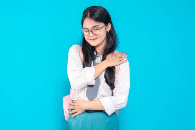 Endonezyalı bir lise öğrencisi, okul üniforması içinde, rahatlatıcı bir pozda kendine sarılırken nazikçe gülümsüyor. Parlak mavi bir arka planda duran bir sırt çantası ve gözlük takıyor..