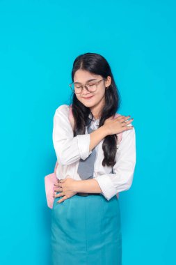 Endonezyalı bir lise öğrencisi, okul üniforması içinde, rahatlatıcı bir pozda kendine sarılırken nazikçe gülümsüyor. Parlak mavi bir arka planda duran bir sırt çantası ve gözlük takıyor..