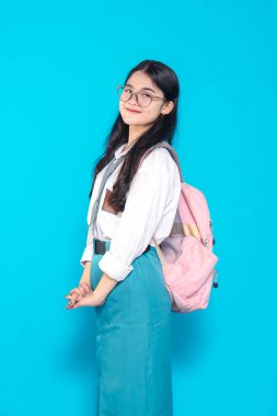 Endonezyalı bir lise öğrencisi, gözlüklü ve sırt çantalı, bir ayağı havada şakacı poz veriyor. Neşeli bir şekilde gülümsüyor, parlak mavi bir arka plana karşı neşeli ve enerjik bir hava yayıyor..