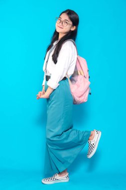 Endonezyalı bir lise öğrencisi, gözlüklü ve sırt çantalı, bir ayağı havada şakacı poz veriyor. Neşeli bir şekilde gülümsüyor, parlak mavi bir arka plana karşı neşeli ve enerjik bir hava yayıyor..