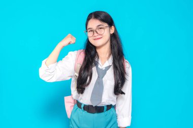 Endonezyalı bir lise öğrencisi gözlüklü ve sırt çantalı güçlü bir jestle kolunu esnetirken güvenle gülümsüyor. Parlak mavi bir arka plana karşı kararlılık ve pozitiflik sergiliyor..