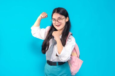 Endonezyalı bir lise öğrencisi, gözlüklü ve sırt çantalı. Heyecanlı ve enerjik bir ifade sergiliyor. Kollarını güçlü bir pozda parlak mavi bir arka plana doğru açıyor..