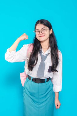 Endonezyalı bir lise öğrencisi gözlüklü ve sırt çantalı güçlü bir jestle kolunu esnetirken güvenle gülümsüyor. Parlak mavi bir arka plana karşı kararlılık ve pozitiflik sergiliyor..