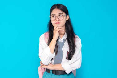 Endonezyalı bir lise öğrencisi gözlüklü ve sırt çantalı, çenesini parlak mavi bir arka planda dinlendirirken düşünceli bir ifade sergiliyor..