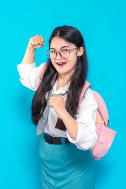 Endonezyalı bir lise öğrencisi, gözlüklü ve sırt çantalı. Heyecanlı ve enerjik bir ifade sergiliyor. Kollarını güçlü bir pozda parlak mavi bir arka plana doğru açıyor..