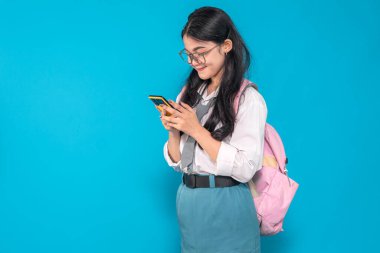 Endonezyalı bir lise öğrencisi gözlüklü ve sırt çantalı akıllı telefonuna bakarken gülümsüyor. Mutlu görünüyor ve parlak mavi bir arkaplanı var..