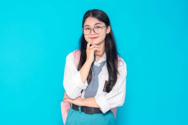 Endonezyalı bir lise öğrencisi gözlüklü ve sırt çantalı, çenesini parlak mavi bir arka planda dinlendirirken düşünceli bir ifade sergiliyor..