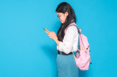 Endonezyalı bir lise öğrencisi gözlüklü ve sırt çantalı akıllı telefonuna bakarken gülümsüyor. Mutlu görünüyor ve parlak mavi bir arkaplanı var..