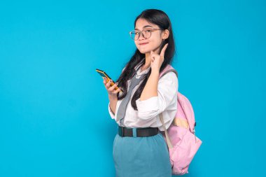 Endonezyalı bir lise öğrencisi gözlüklü ve sırt çantalı, akıllı bir telefonu tutarken özgüvenle gülümseyen ve parlak mavi bir arka plana karşı harika bir fikri varmış gibi parmağını kaldıran..